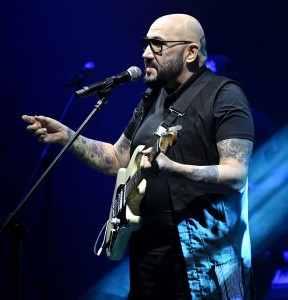 Artysta stojący na scenie z gitarą i śpiewający do stojącego przed nim mikrofonu. Prawa dłoń podniesiona. Na obu przedramionach widoczne tatuaże. Z tytułu i częściowo na postaci smuga światła. Widok z perspektywy lewej strony artysty. . Kliknij, aby powiększyć zdjęcie.